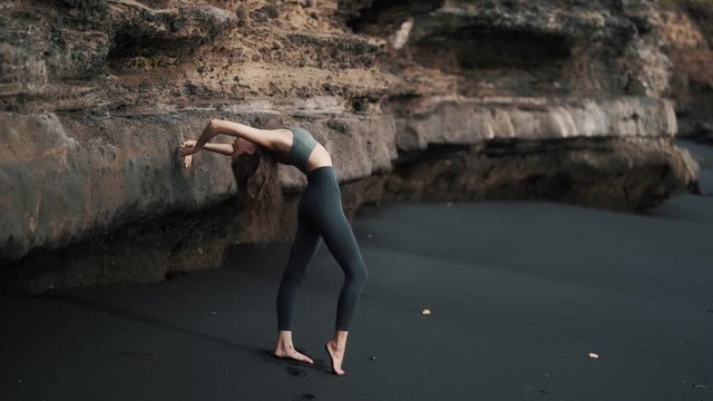 Woman doing stretching exercises on black sand beach near cliff, slow motion