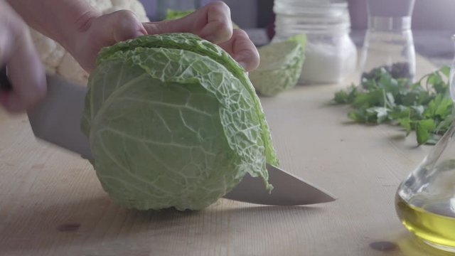 Close-up bottom view of a woman's hands cutting a savoy cabbage in two with a knife on a wooden chopping board with vegetables, oil, salt, pepper and parsley on the blurred background slog3 ungraded