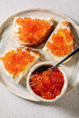 Red salmon caviar in bowl and on wheat bread, served on ceramic plate over grey spotted background. Flat lay, space