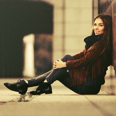 Happy young fashion woman in leather jacket sitting on sidewalk