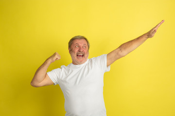Successful winner's gesture. Caucasian man portrait isolated on yellow studio background. Beautiful male model in white shirt posing. Concept of human emotions, facial expression, sales, ad. Copyspace