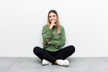 Young caucasian woman sitting on the floor smiling happy and confident, touching chin with hand.