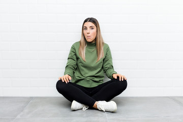 Young caucasian woman sitting on the floor shrugs shoulders and open eyes confused.