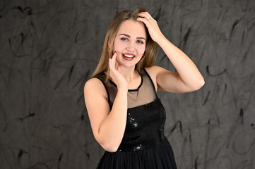 Fototapeta premium Universal concept of horizontal female portrait on gray alternative background. A photo of a pretty smiling girl with long hair and excellent make-up in a black dress stands in different poses.