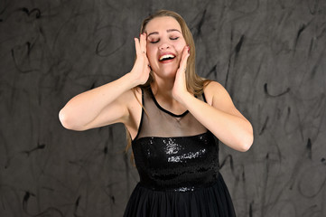 Fototapeta premium A photo of a pretty smiling girl with long hair and excellent make-up in a black dress stands in different poses. Universal concept of horizontal female portrait on gray alternative background.