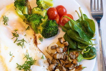Proper nutrition, healthy eating breakfast of eggs, broccoli, mushrooms, garlic, spinach, dill and small tomatoes close up on white plate with fork lying near. Diet balanced organic food breakfast