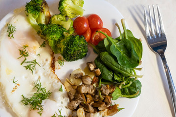 Proper nutrition, healthy eating breakfast of eggs, broccoli, mushrooms, garlic, spinach, dill and small tomatoes close up on white plate with fork lying near. Diet balanced organic food breakfast