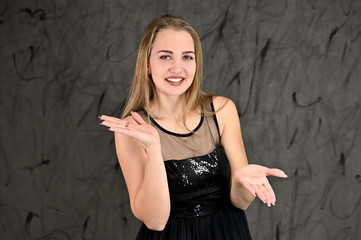 Fototapeta na wymiar A photo of a pretty smiling girl with long hair and excellent make-up in a black dress stands in different poses. Universal concept of horizontal female portrait on gray alternative background.