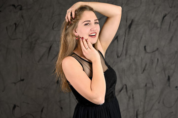 Fototapeta premium A photo of a pretty smiling girl with long hair and excellent make-up in a black dress stands in different poses. Universal concept of horizontal female portrait on gray alternative background.