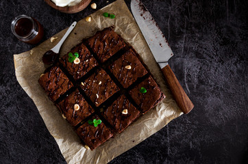 Homemade chocolate brownie dessert with hazelnuts, chocolate glaze and whipped cream on baking paper on a dark background. Top view. Horizontal orientation. Copy space.