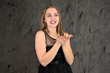 A photo of a pretty smiling girl with long hair and excellent make-up in a black dress stands in different poses. Universal concept of horizontal female portrait on gray alternative background.