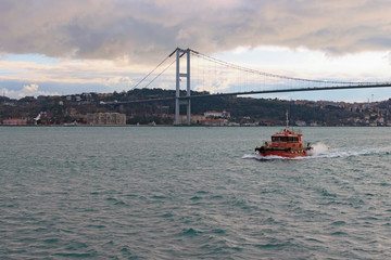 Landscape view of The Bosporus (Bosphorus or Strait of Istanbul) on a cloudy day. Red motorboat cruising in The Bosporus. The Bosphorus Bridge (15 July Martyrs Bridge, First Bridge) in the background