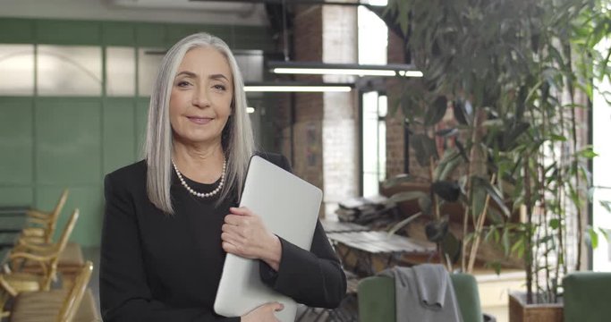 Matured businesswoman standing in modern working space and holding laptop. Successful midle aged woman looking to camera and smiling. Portraits. Concept of business and work.