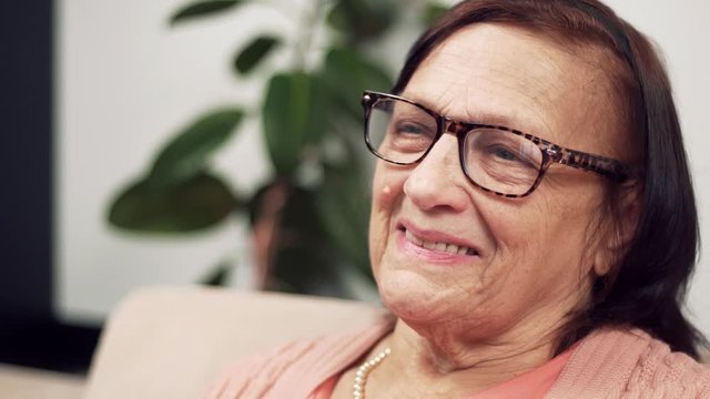 Elderly Woman Smiling Portrait Close-up. Mature Woman In Glasses Portrait Close Up.Happy Mature Senior Cheerful Aged Retired Grandmother Sitting On Sofa Looking At Camera.Positive Old Female Smiling. 
