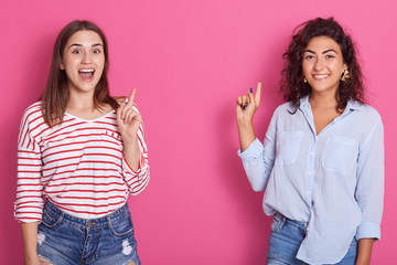 Image of smiling adorable beautiful brunettes making gestures, raising forefingers, showing direction, looking directly at camera, being in good mood. Copyspace for advertisement. Emotions concept.