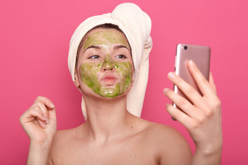 Horizontal shot of young beautiful girl with dried face mask on her face using her smart phone for...