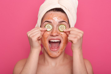 Studio shot of satisfaited woman applies slices of cucumber on her eyes and having scrabmask on face, has happy expresion, opens mouth widelly, doing beauty procedures at home, wears white towel.