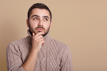Obraz premium Close up portrait of handsome bearded man posing in knitted warm sweater, isolated over beige background, keeping hand on chin, looking aside. Copy space for advrertisment or pronotional text.