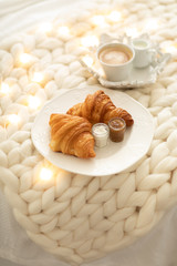 Fresh croissants with jams and americano with milk on knitted white wool blanket and luminous garlands. Cozy winter morning at home. Scandinavian bedroom.