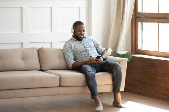 Handsome african guy sitting on couch listens music holding smartphone