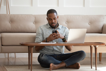 African guy sits cross-legged on carpet using smartphone