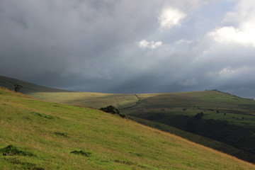 Landscape photo of steep hills with hedge fields covered by lush green grass. Rays of sun are falling through the clouds to the valley. Dartmoor National Park, next to the Meldon Reservoir, UK