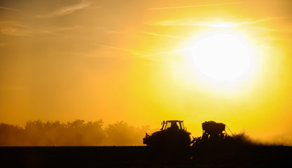 Fototapeta na wymiar Silhouette of a tractor sowing seeds in a field in a cloud of dust against the background of the setting sun.