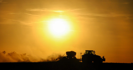 Silhouette of a tractor sowing seeds in a field in a cloud of dust against the background of the...