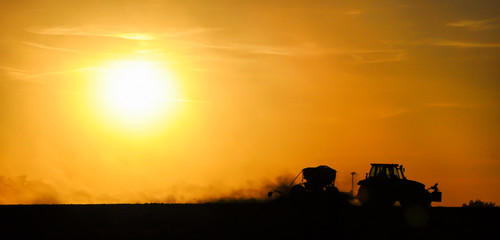 Silhouette of a tractor sowing seeds in a field in a cloud of dust against the background of the...