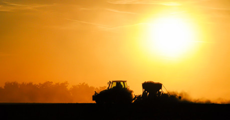 Fototapeta premium Silhouette of a tractor sowing seeds in a field in a cloud of dust against the background of the setting sun.