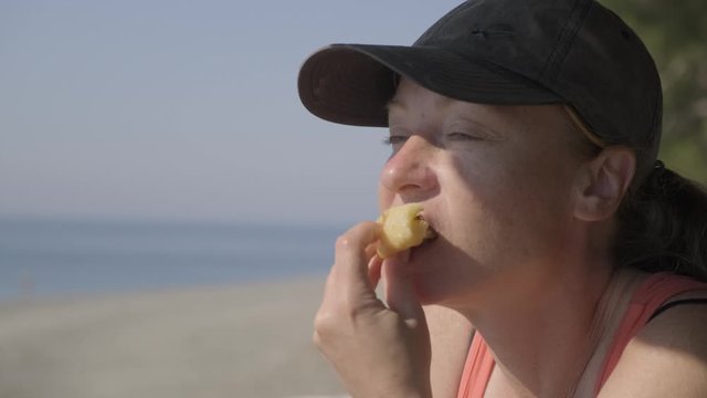 Portrait of adult woman in baseball cap happily eating pie on background of seashore.