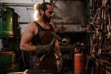 Fototapeta na wymiar Brutal muscular artisan blacksmith standing in the workshop holding a hammer in his hand in front of a wall of tools
