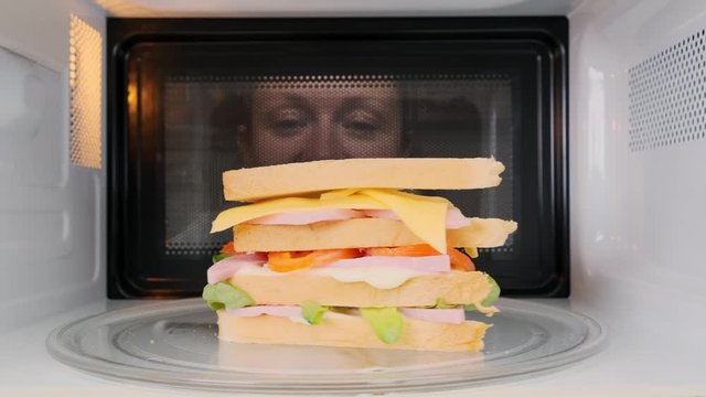 big sandwich is heated in microwave oven when woman watches cooking process through glass of door.