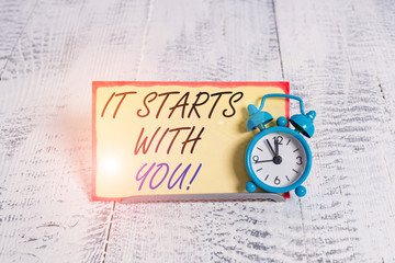 Writing note showing It Starts With You. Business concept for Motivation for starting doing something Have discipline Alarm clock tilted above buffer wire in front of notepaper