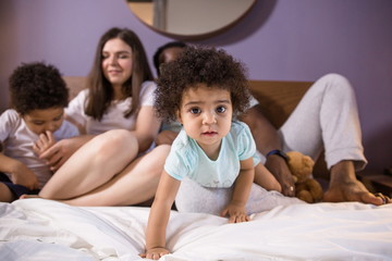 Happy ethnic family lying on the bed in the bedroom. Little sister in the foreground crawling forward