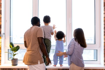 Fototapeta premium Happy ethnic family with two children looking out the window standing in the apartment