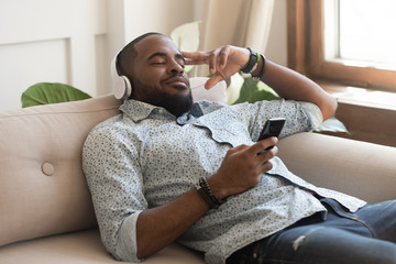 African guy holding smartphone closed eyes listen music on headphones