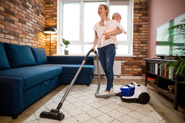 Fototapeta na wymiar Young beautiful mother holding a newborn baby in her arms, cleaning the floors with a vacuum cleaner in the apartment