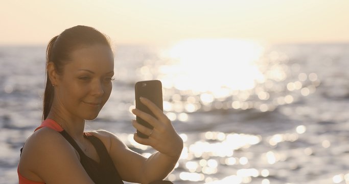 Young girl with long pony tail and a beautiful athletic body takes a selfie on the front camera on the background of the sea in the sunrise