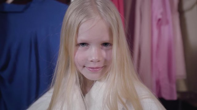 Close-up face of beautiful Caucasian girl with grey eyes and long blond hair looking at camera and smiling. Portrait of charming child in white bathrobe posing at home.