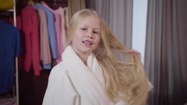 Camera approaching to charming Caucasian girl drying hair with towel. Portrait of smiling child resting in her room on weekends. Joy, hygiene, leisure.