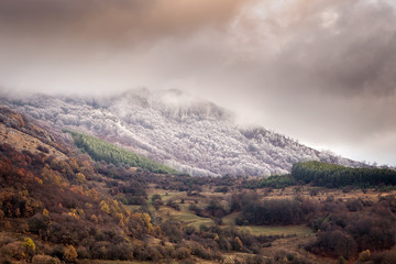 Amazing, moody, rocky summit covered in frost and fog, autumn colors of the forest trees below on the transition period between autumn and winter