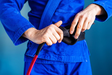 Close up on midsection of the bjj brazilian jiu jitsu black belt hands holding and tie around the waist of an athlete fighter wearing blue kimono gi standing in front of the wall