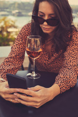 Beautiful elegant woman drinking wine and using cellphone outdoors.
