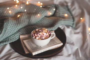 Cup of hot chocolate with marshmallow staying on open paper books with knitted sweater over glowing xmas lights closeup. Good morning. Breakfast. Autumn season.