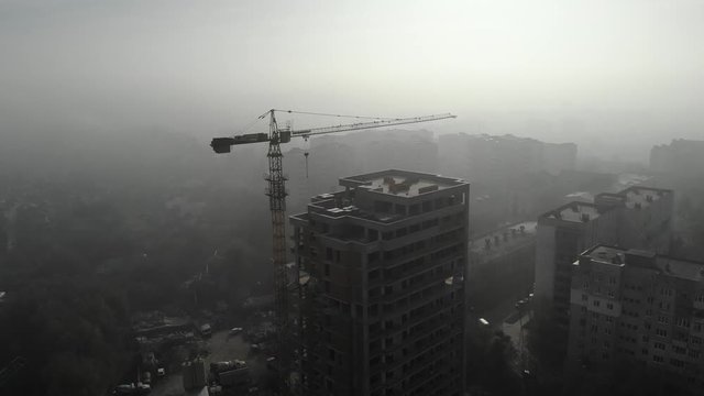 Birds eye view on tower crane in fog standing next to residential building. Flying over the construction site. Drone real time footage.