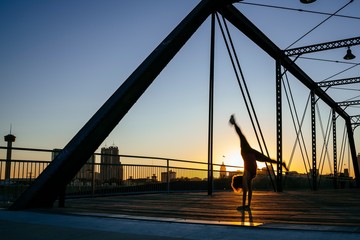 Woman in fitness attire doing hand stand on pedestrian bridge in city at sunset