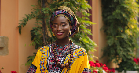 Portrait shot of the beautiful and happy Afrcan woman in the traditional outfit standing in the cozy courtyard with flowers and smiling to the camera. Outdoor.