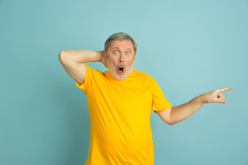 Fototapeta premium Shpcked pointing at side. Caucasian man portrait isolated on blue studio background. Beautiful male model in yellow shirt posing. Concept of human emotions, facial expression, sales, ad. Copyspace.