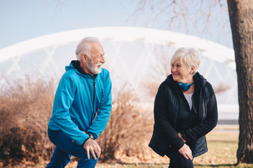 An older man and an older woman stretching their knees in the park. The woman is wearing blue headphones around her neck.